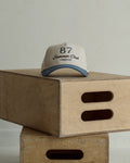 Beige and light blue cap with '87 Summer Club Company' text on a wooden box against a light gray background