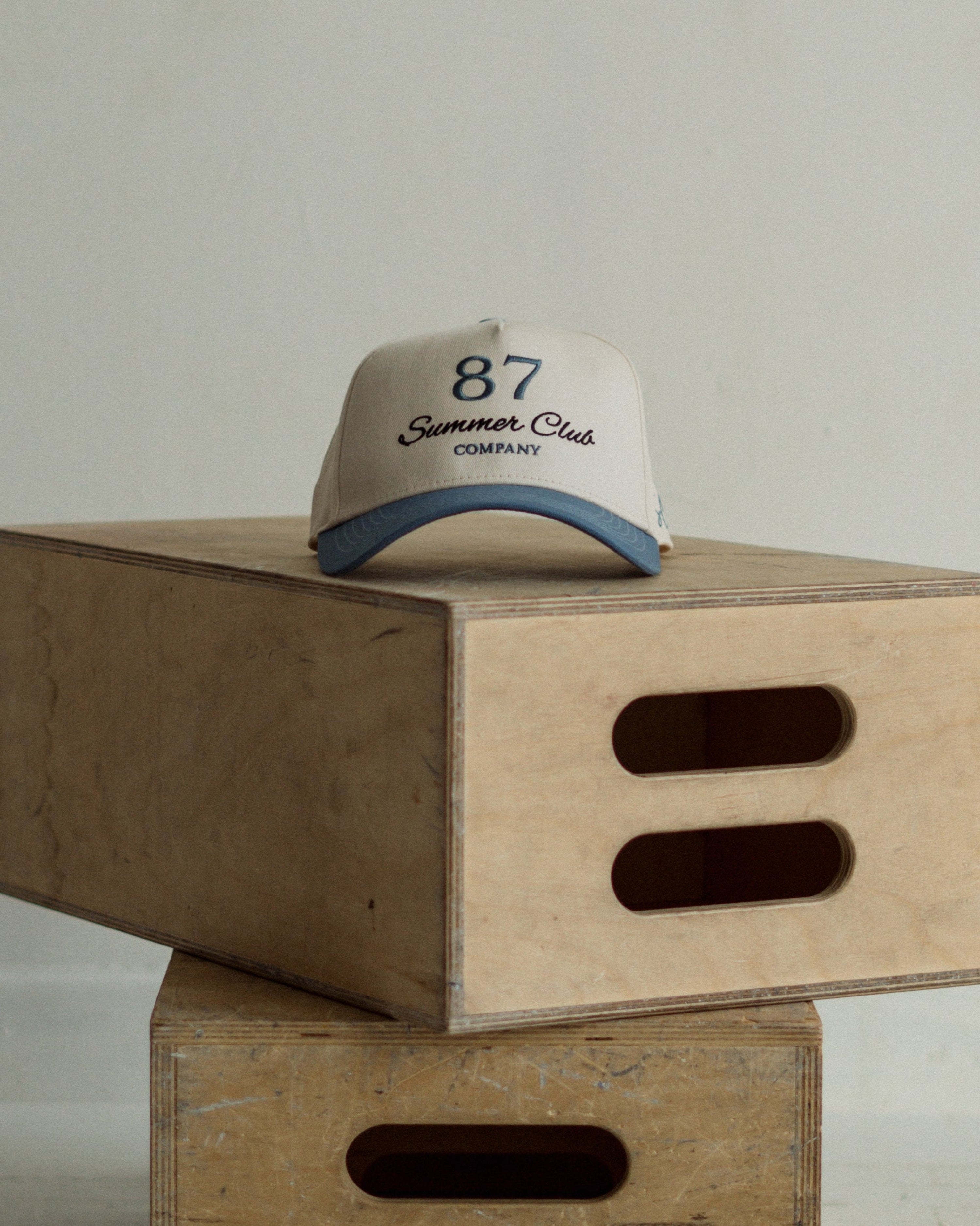 Beige and light blue cap with '87 Summer Club Company' text on a wooden box against a light gray background